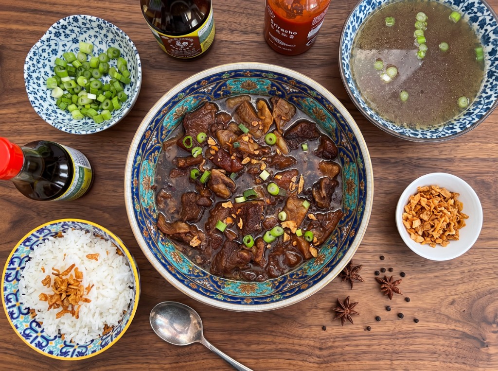 Beef pares set on a wooden table: glossy braised beef in a blue-and-gold bowl topped with green onions and fried garlic, garlic rice, a bowl of clear beef soup, extra fried garlic, and star anise pods on the side