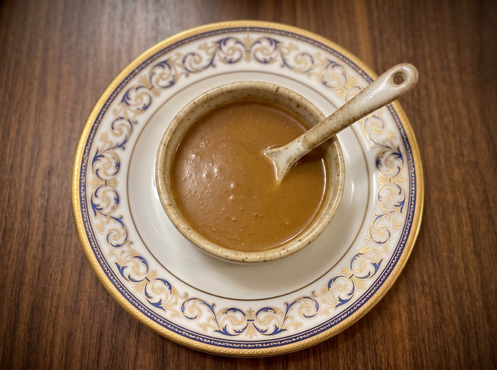 Glossy brown Chickenjoy gravy in a rustic speckled ceramic bowl with a matching spoon, set on an ornate blue-and-gold plate—smooth, lump-free sheen from potato starch