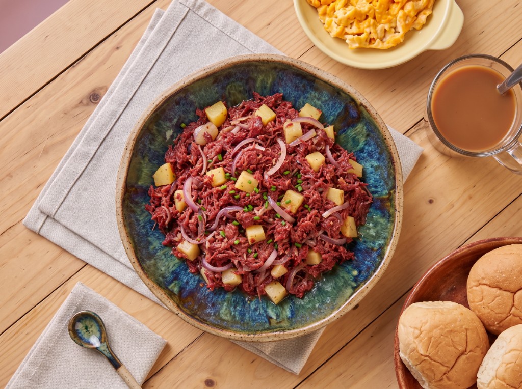 Corned beef ginisa in a rustic blue-green bowl: shredded pink corned beef with yellow potato cubes, red onion rings, and chives, served with pandesal, scrambled eggs, and coffee