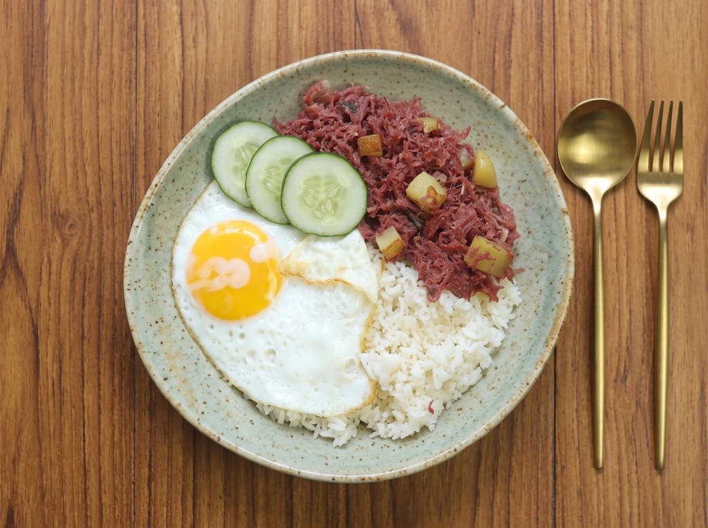 Cornsilog in a speckled bowl: shredded corned beef with fried potato cubes, white garlic rice, a sunny-side-up egg, and cucumber slices, gold spoon and fork on wood