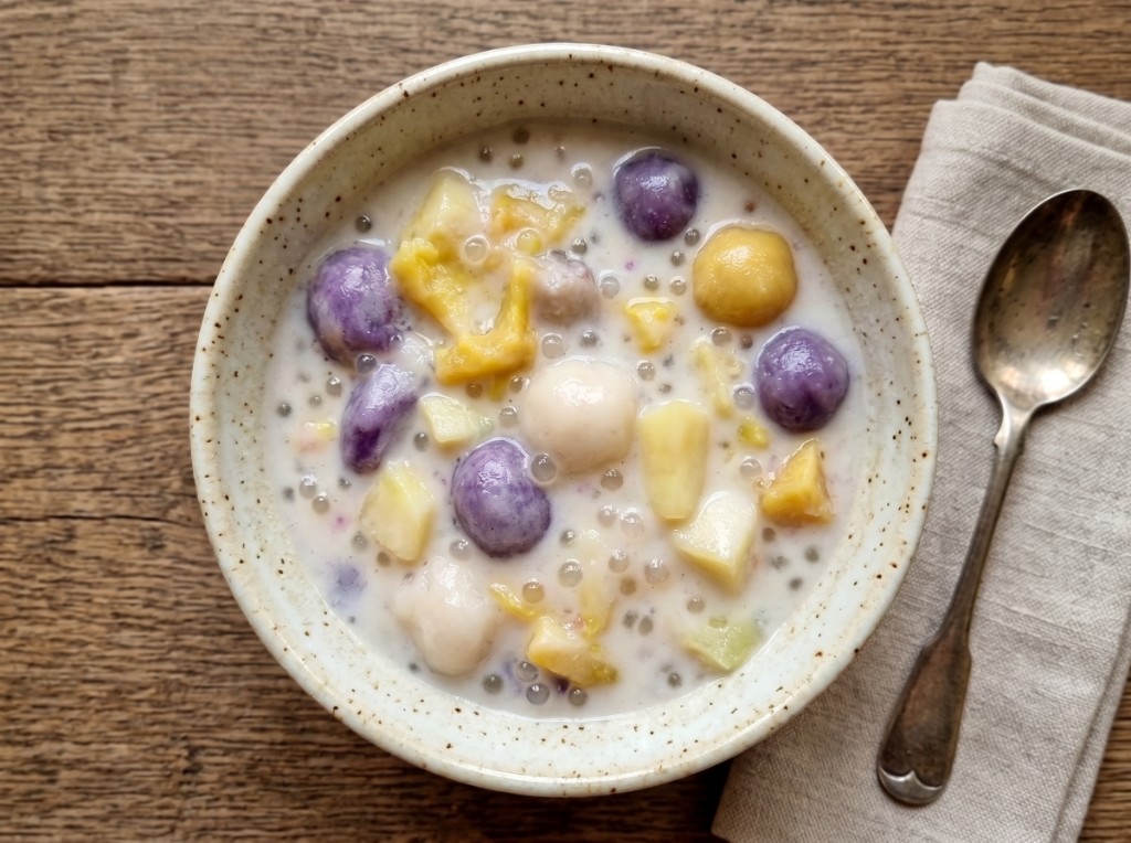 Bowl of ginataang bilo-bilo on a wooden table: creamy coconut milk packed with glossy purple ube rice balls, white bilo-bilo, yellow jackfruit, kamote cubes, and tiny translucent sago pearls