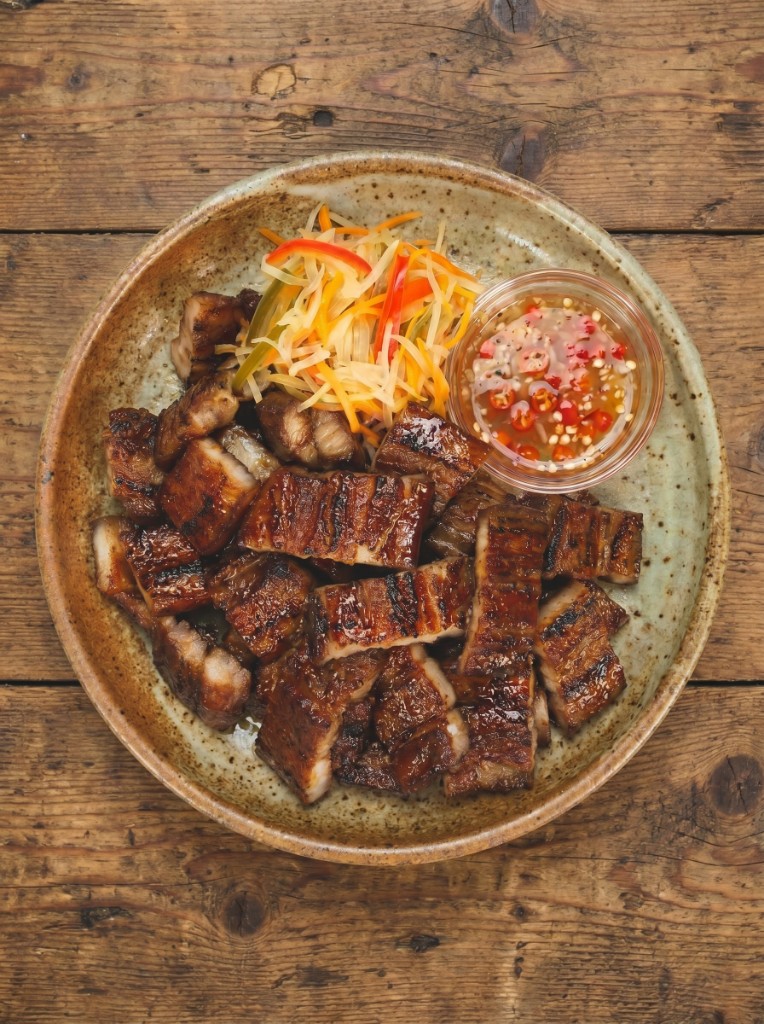 Inihaw na liempo on a rustic speckled plate, top-down: bite-sized grilled pork belly slices with char marks and glossy soy glaze, atchara on the side, and a small bowl of vinegar sawsawan with red chili on a wooden table