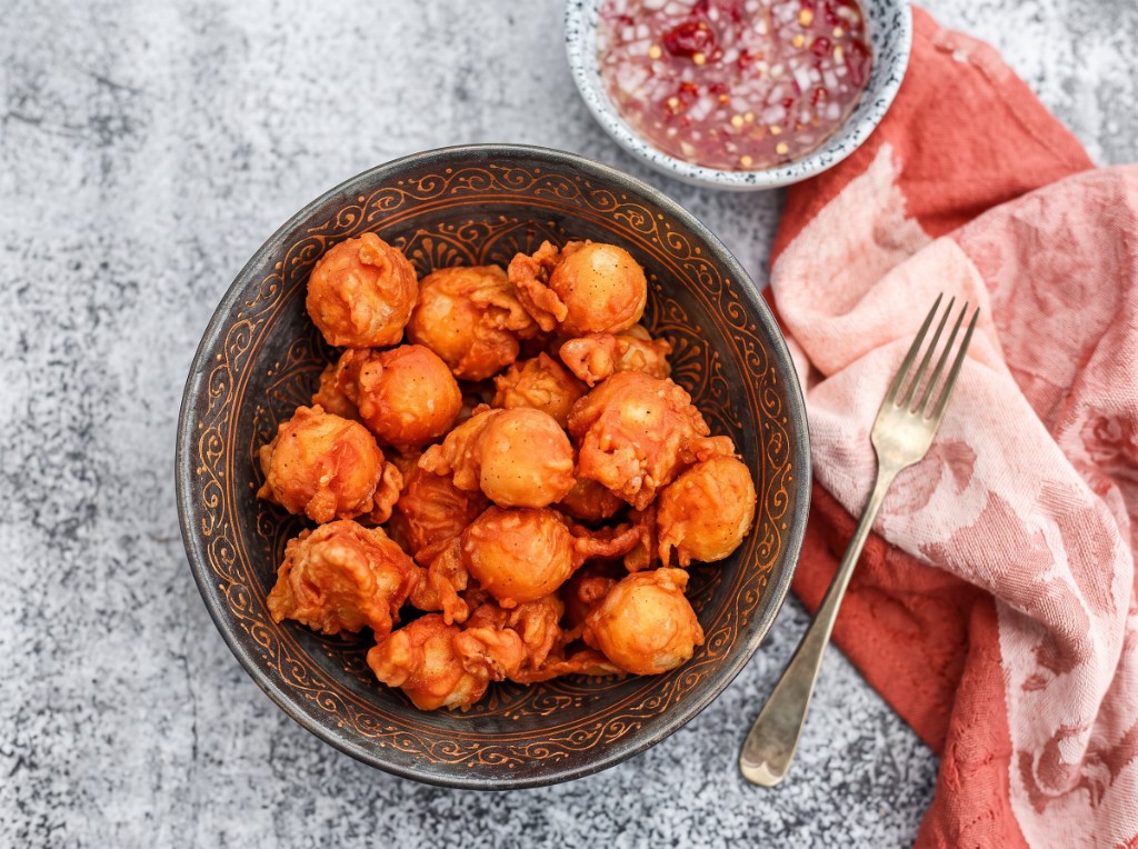 Kwek-kwek piled in a dark patterned bowl: crispy golden-orange battered quail eggs with craggy fried batter, vinegar dipping sauce with minced onion and red chili, pink napkin and fork