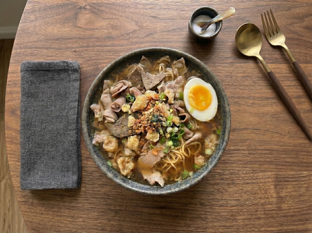 La Paz batchoy in a dark ceramic bowl: clear amber broth with thin yellow miki, pork slices, liver, pork intestines, piled toasted garlic and crushed chicharon, green onions, and a halved soft-boiled egg with jammy orange yolk
