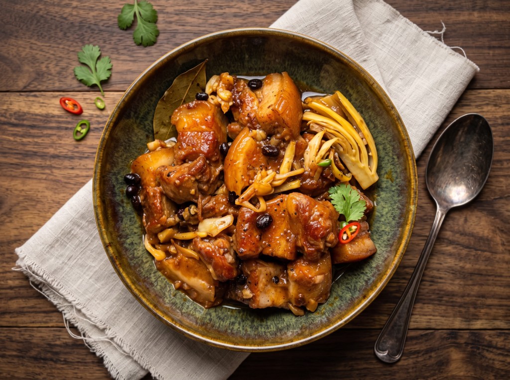 Pork humba in a rustic dark bowl: glossy braised pork belly in thick sweet-savory sauce with tausi, banana blossoms, a bay leaf, cilantro, and sliced red and green chilies