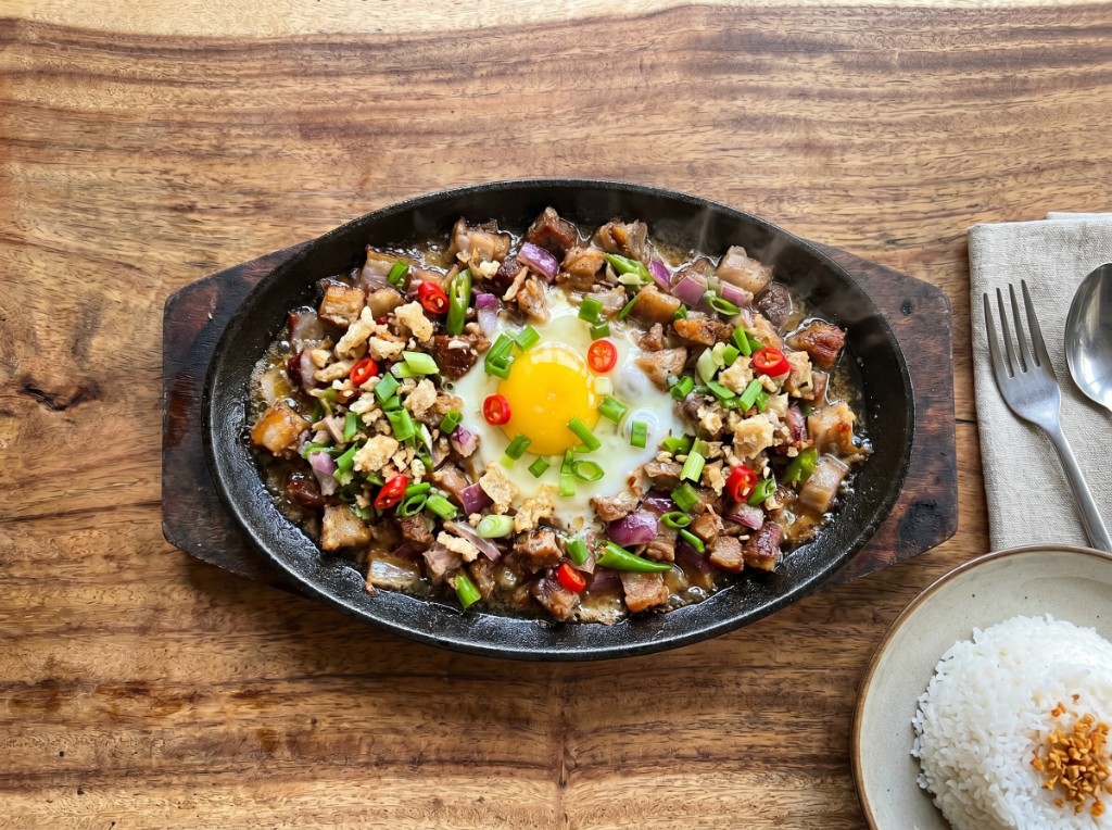 Sizzling pork sisig on a cast iron plate with egg, onions, chilies, chicharon, and garlic rice on the side
