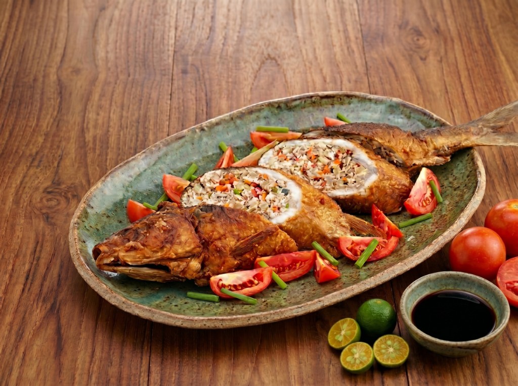 Relyenong bangus on a rustic oval platter: golden pan-fried stuffed milkfish sliced diagonally to show flaked filling with carrots and peas, tomato wedges and green onions around the fish, calamansi halves and soy dipping sauce on the side
