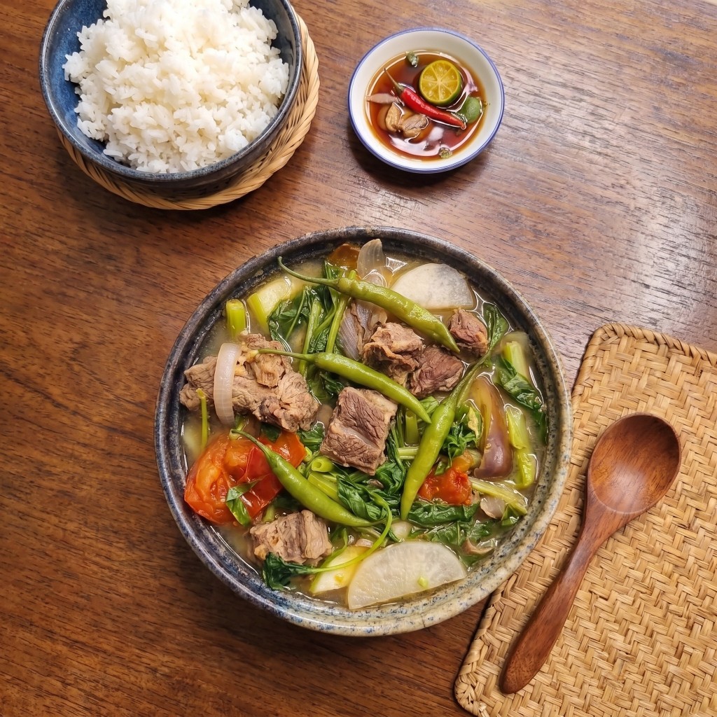 Sinigang na baka table: rustic speckled bowl of beef sour soup with kangkong, labanos, eggplant and siling haba, steamed rice, patis sawsawan with calamansi and red chili, wooden spoon