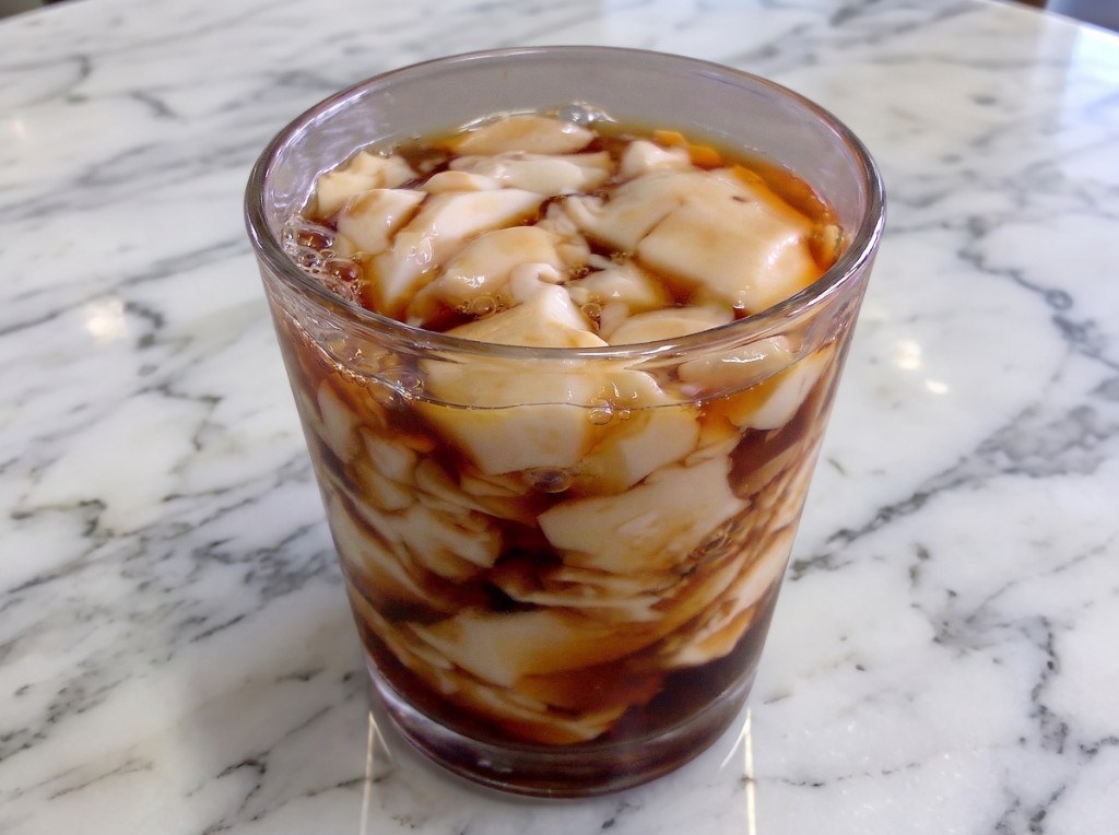 Glass of taho with silken tofu chunks in dark amber arnibal syrup on a marble surface