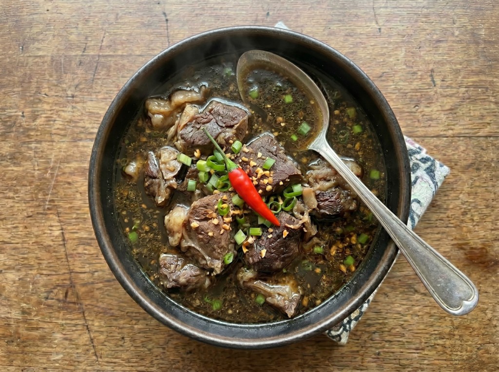 Tyula itum in a dark ceramic bowl on a wooden table: deep black glossy broth with large tender beef chunks, fried garlic, green onions, and a whole red siling labuyo on top; steam rising